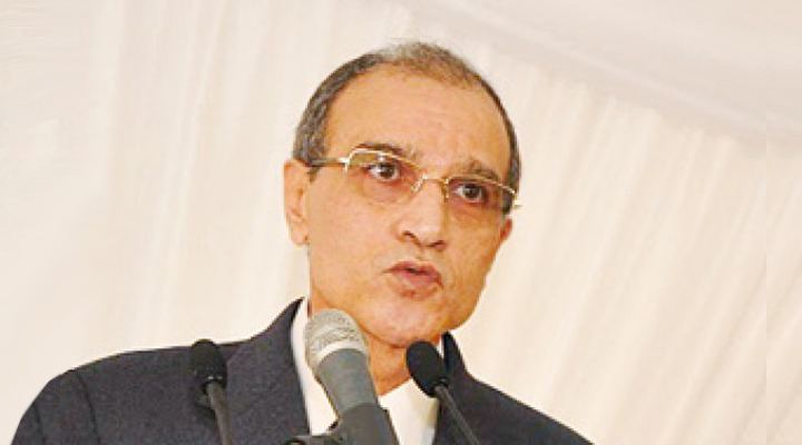 Ali Wakrim, wearing glasses and a dark suit, speaking into microphones at a lectern in front of a pale draped backdrop.
